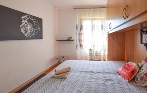 Ein Schlafzimmer mit Bett, einem kleinen Tisch, einer Bücherablage, einem Gemälde und einem Fenster mit Vorhängen.
