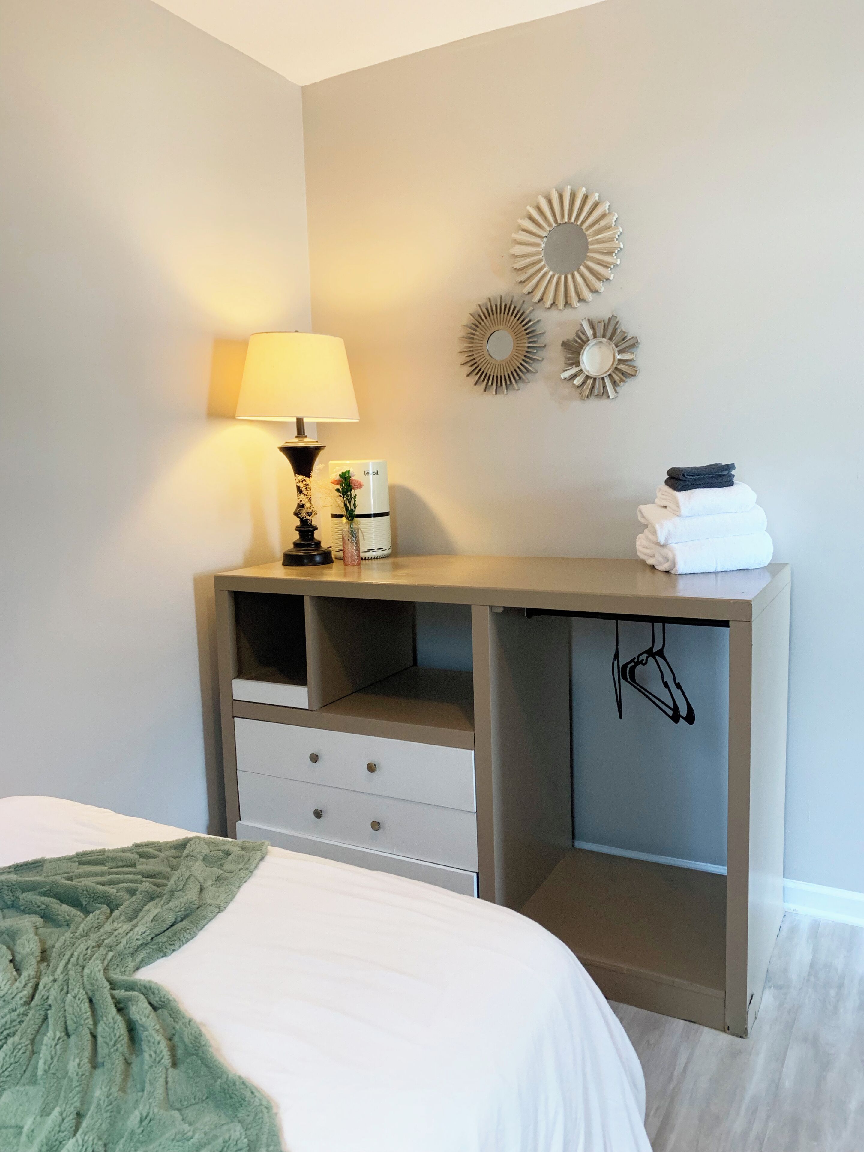 Een slaapkamer met een bed, een nachtkastje met een lamp, een spiegel en een wandplank met handdoeken.