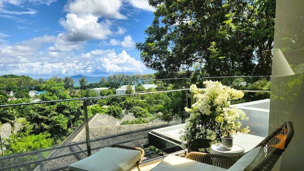 A balcony with two wicker chairs and a table, overlooking a lush garden and distant buildings. - Sea View the tree 1BR Condo 500m Walk 2 BeachC260 (Tambon Rawai)