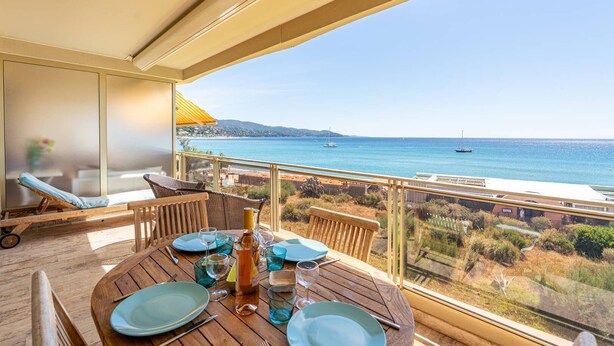 Ein Balkon mit Esstisch, der Blick auf die Küste mit Booten und einem Strand bietet.
