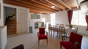 A spacious room with wooden beams, a dining area with red chairs, a kitchen with a dishwasher, and a staircase.