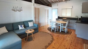 Un espace de vie compact comprenant un canapé, une petite table ronde, un coin repas avec des chaises, un coin cuisine et un plafond avec des poutres apparentes.