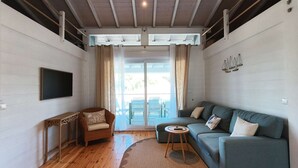 Un salon comprenant un canapé, un fauteuil, un téléviseur et donnant sur l’extérieur.