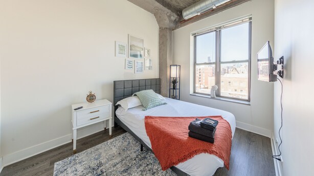 A modern bedroom with a large bed, a nightstand, a bedside table, a window with a city view, and a ceiling with exposed ductwork.