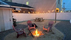 Un patio trasero con un pozo de fuego, cuatro sillas Adirondack, una mesa de picnic y luces colgantes.