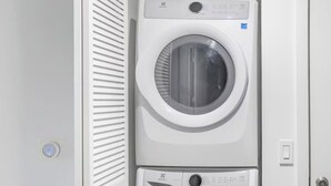 Un lavadero pequeño y blanco con dos lavadoras apiladas, una puerta con celosía y un interruptor de luz en la pared.