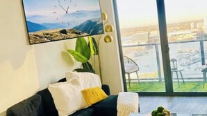 Una sala moderna con un sofá negro, una mesa de centro con objetos decorativos, un ventanal con vista a la ciudad y un reloj montado en la pared.