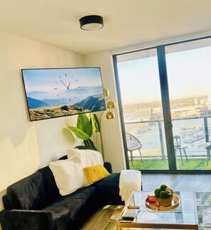 Ein modernes Wohnzimmer mit einer schwarzen Couch, einem Couchtisch mit Dekoration, einem großen Fenster mit Blick auf die Stadt und einer Wanduhr.