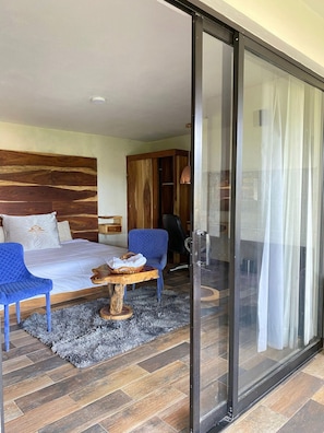 A bedroom with a bed, a blue chair, a wooden side table, and a glass door leading to a balcony.