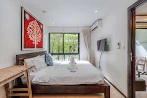 Ein Schlafzimmer mit Bett, einem Holznachttisch, einem Fernseher und einem Fenster mit Blick ins Grüne.