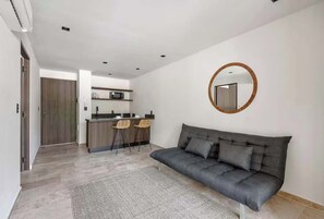 A modern living room with a grey sofa, a round mirror, and a kitchen in the background. - Chic Jungle 1BR: Balcony Priv Pool, Steam & Gym (Tulum)