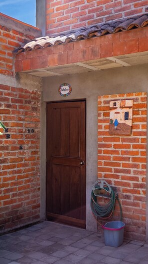 A brick building with a wooden door, a sign reading 'Caballeros,' a small wall-mounted artwork, and a hose coiled on the ground.