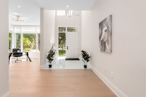 A spacious room with a wooden floor, a black office chair, a desk, two potted plants, and a large window with a view of greenery.