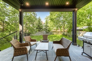 Un patio exterior con mobiliario de mimbre, una parrilla y vistas a un jardín trasero.