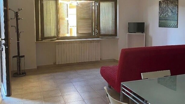 Ein Zimmer mit einem roten Sofa, einem Glastisch, einem Heizkörper, einem Fenster mit Vorhängen, einem Kleiderständer und einem Fernseher auf einem Schrank.