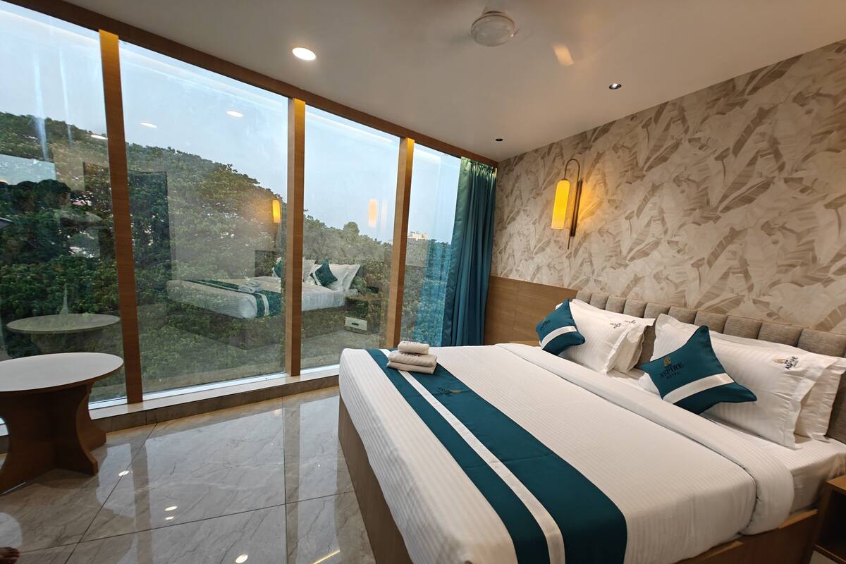 Ein Hotelzimmer mit einem großen Bett, einem runden Tisch und Blick auf Grün hinter raumhohen Fenstern.