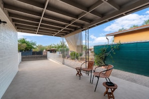 Un patio techado con piso de concreto, dos sillas de mimbre, una mesita redonda y una cerca verde al fondo.