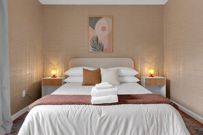 Habitación de hotel con cama, dos mesitas de noche con lámparas, un cuadro colgado en la pared y una alfombra estampada.