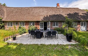 Une maison en briques avec un toit de chaume, une terrasse avec une table et des chaises, et un jardin.