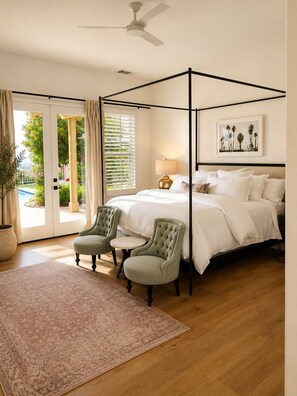 Een hemelbed met nachtkastje, twee fauteuils, een klein rond tafeltje, een vloerlamp en een groot raam met jaloezieën.