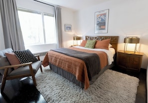 A bedroom with a bed, a chair, a nightstand, a lamp, and a window.