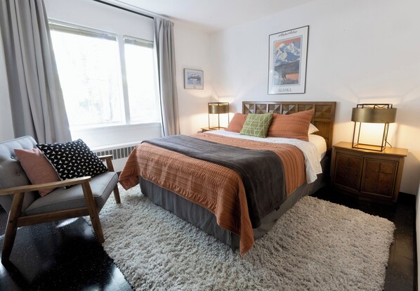 Une chambre Ă coucher avec un lit, une chaise, une table de chevet, une lampe et une fenĂȘtre.