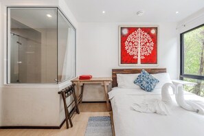 Ein modernes Hotelzimmer mit Duschkabine aus Glas, einem Bett mit weißer Bettwäsche, einem roten abstrakten Gemälde und Blick auf Grün durch ein großes Fenster.