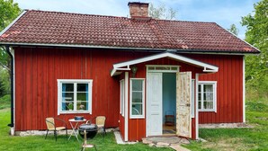 Una casa de madera roja con techo de tejas, una puerta principal y un pequeño porche con sillas y una parrilla.