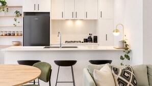 Cozinha moderna com armários brancos, geladeira de aço inoxidável, fogão a gás e máquina de café. Possui dois banquetas de bar e uma mesa de jantar redonda.