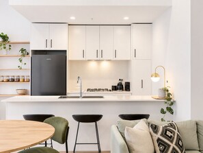 A modern kitchen with white cabinets, a stainless steel refrigerator, a gas stove, and a coffee maker. There are two bar stools and a round dining table. - Moonee Ponds Luxe by Ready Set Stay (Moonee Ponds)