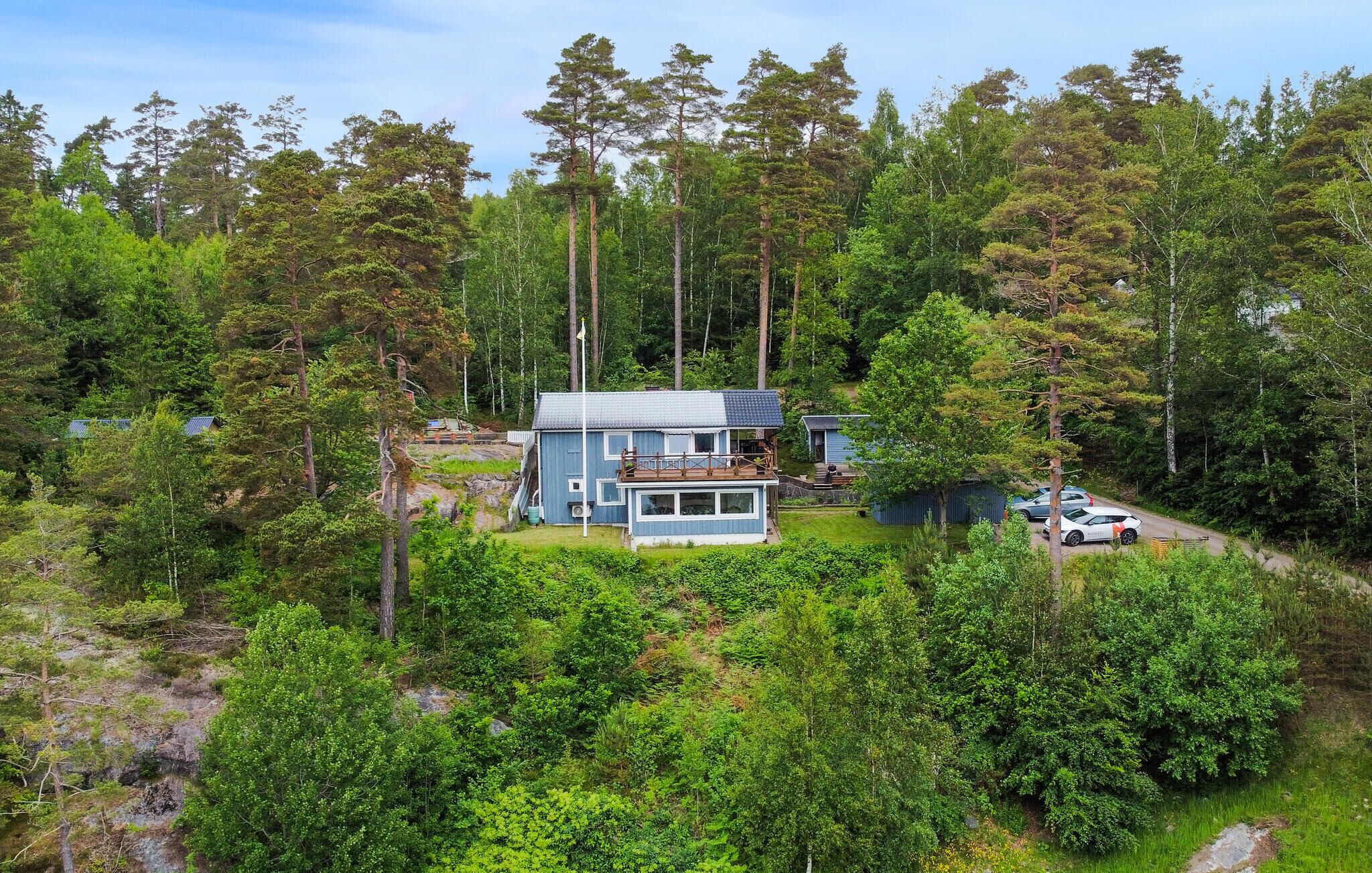 En modern stuga i två våningar, omgiven av tät skog, med en synlig veranda och en liten tillbyggnad.