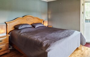A bedroom with a wooden headboard, a dark quilted bedspread, two pillows, a nightstand with a lamp, and a window with a view of trees. - 3 bedroom amazing home in Uddevalla (Uddevalla)