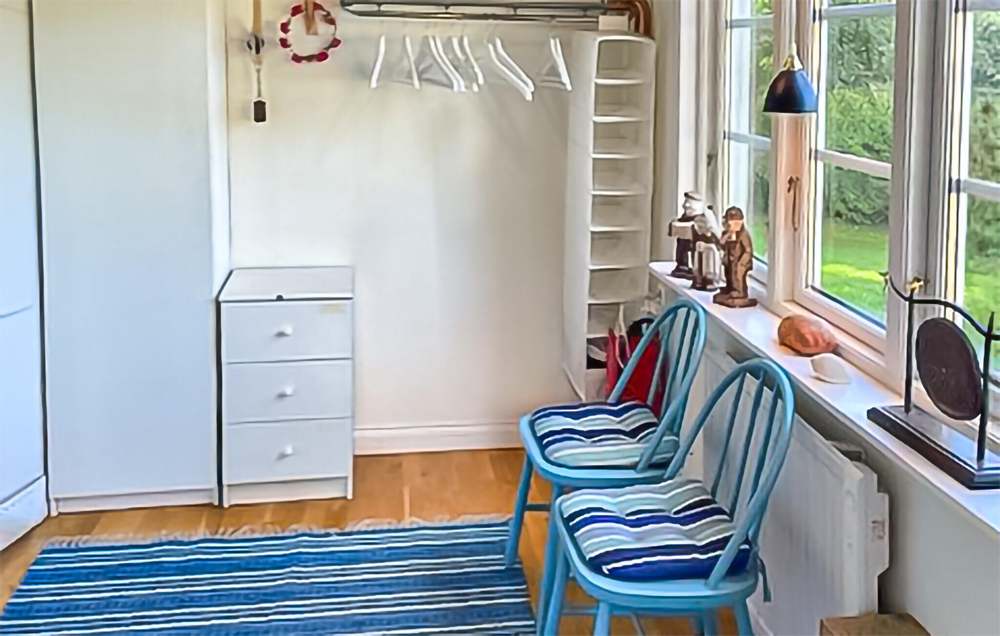 A room with a radiator, a small white cabinet, a blue striped rug, two blue chairs, and a window with a view of greenery.