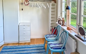 Ein Zimmer mit Heizkörper, einem kleinen weißen Schrank, einem blauen Teppich mit Streifen und zwei blauen Stühlen. Ein Fenster bietet Blick auf Grünflächen.