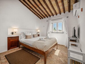 A bedroom with a wooden bed, bedside lamps, a window with curtains, a TV on a stand, and a herringbone-patterned floor. - Quinta da Luz, Rustic Villa with Pool and Sea Views (Luz, Lagos)