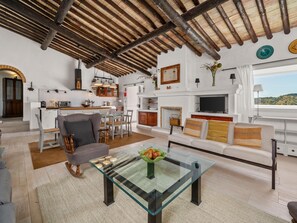 A modern living room with a glass coffee table, a fireplace, a TV, and a kitchen area with wooden beams. - Quinta da Luz, Rustic Villa with Pool and Sea Views (Luz, Lagos)