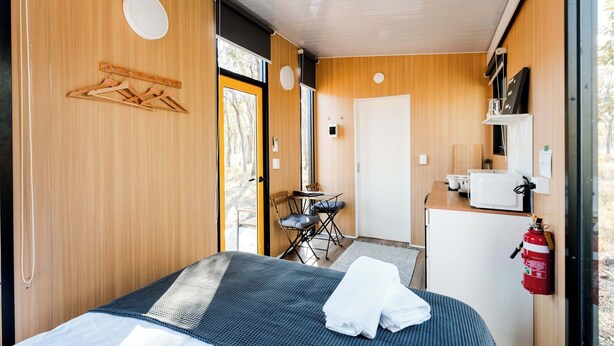 Quarto compacto com cama, uma mesinha, uma cadeira, micro-ondas e extintor de incêndio.