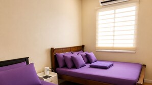 Quarto com duas camas, uma mesinha de cabeceira, um abajur e uma janela com persianas.