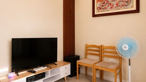 Uma sala de estar com televisão de tela plana, cadeiras de madeira, um ventilador de pé e uma obra de arte emoldurada na parede.