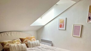 Une chambre avec un plafond en pente, un lit recouvert de plusieurs coussins, un radiateur et des tableaux encadrés accrochés au mur.