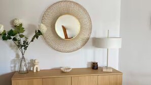 Un meuble en bois avec un miroir rond, un vase de fleurs blanches, une lampe et un panier tressé.