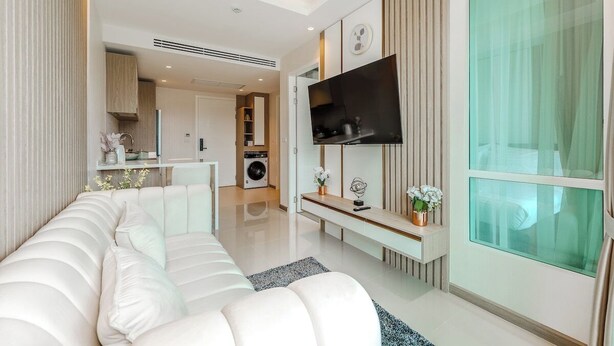 A modern living room with a sofa, a TV mounted on the wall, and a kitchen in the background. - CoWorking Pool-Gym-Game Room+Cinema the city C220 (Tambon Wichit)