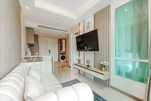 A modern living room with a sofa, a TV mounted on the wall, and a kitchen in the background. - CoWorking Pool-Gym-Game Room+Cinema the city C220 (Tambon Wichit)