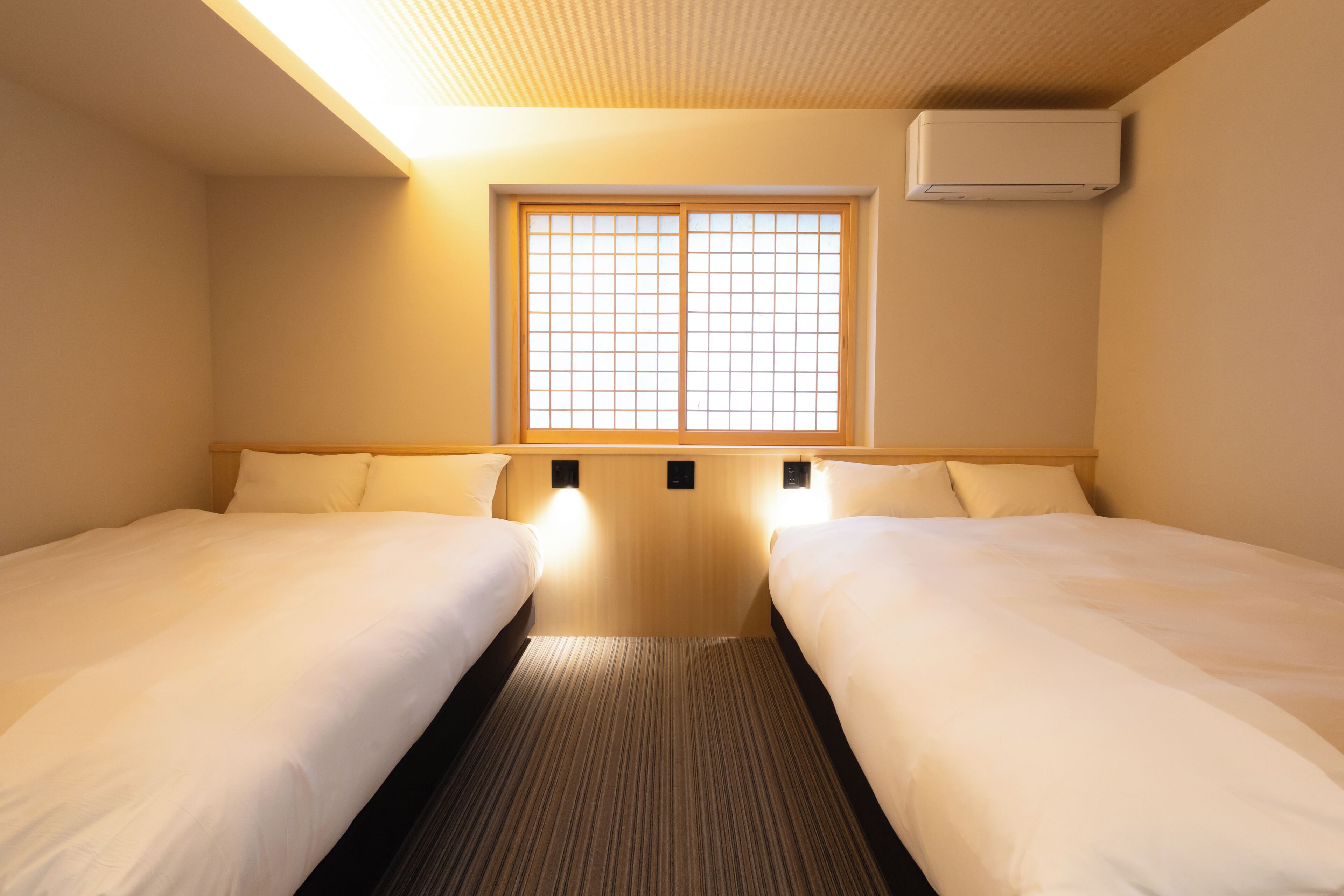 A hotel room with two beds, a window with a grid pattern, and an air conditioning unit on the wall.