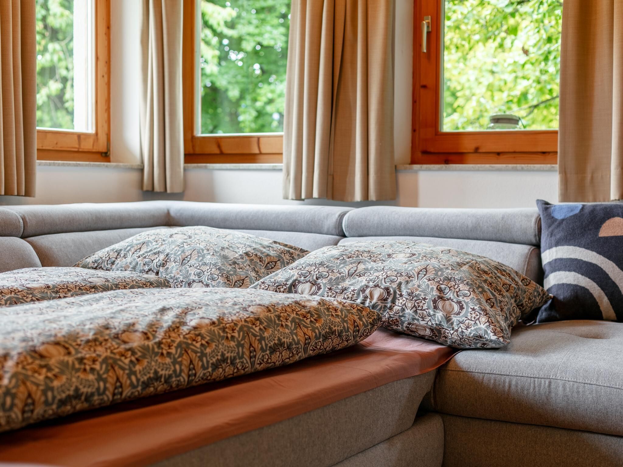 Un canapé avec des coussins à motifs et un banc en bois, de grandes fenêtres avec des rideaux et une vue sur la verdure à l’extérieur.