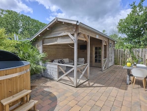 Een houten terras buiten met een overdekte zithoek, een barbecue en een tafel met stoelen.
