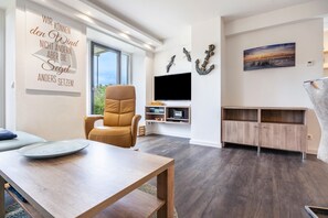 Een moderne woonkamer met een groot raam, een leren fauteuil, een houten tafel, een flatscreen televisie en een kast.