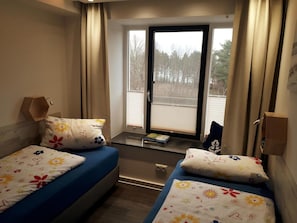 Et værelse med to senge, et vindue med skodder, et lille bord og en lampe på væggen.