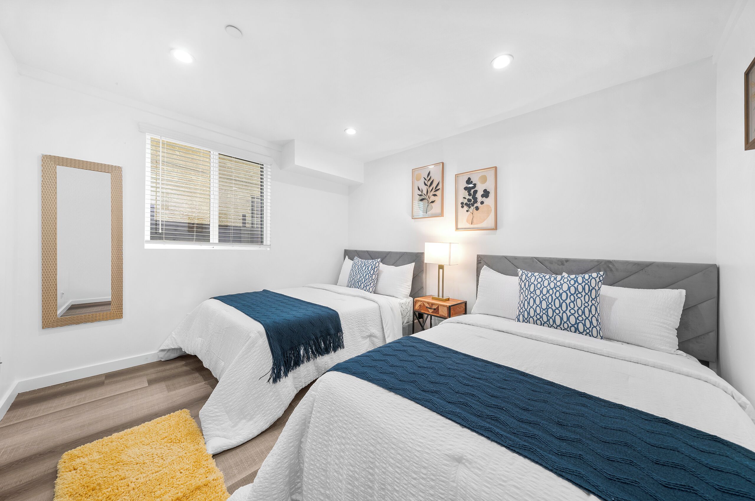 A hotel room with two beds, a large window with blinds, a mirror, and two wall-mounted artworks.