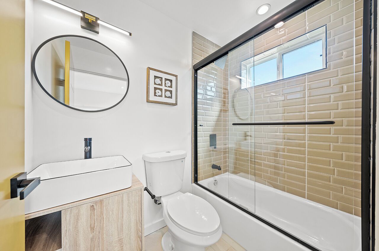 A modern bathroom with a white toilet, a glass-enclosed shower, a round mirror, and a white sink.